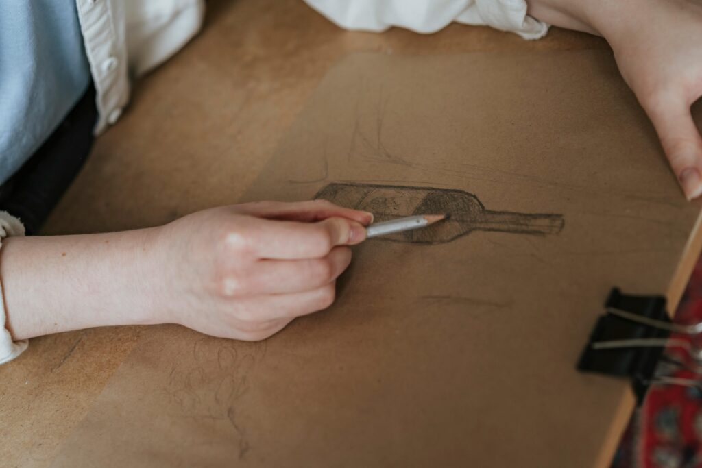 A person sketching a wine bottle on paper with a pencil. Artistic process in a creative setting.